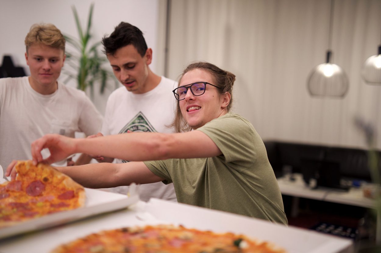 Ein Foto von der Gaming Night im Büro, Elias lacht in die Kamera während er sich ein Stück Pizza nimmt