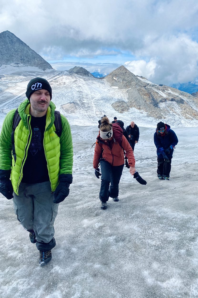 Chris, Anna und der Rest vom Team, laufen mit Winterjacken im Schnee und man sieht Berge im Hintergrund